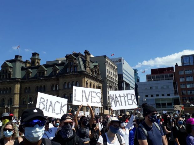 Protestors gather on Parliament Hill with signs raised above their head.