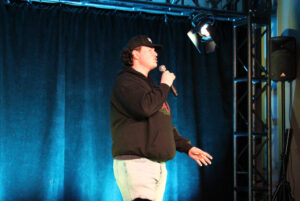 Xander Wynberg, a white man in a CKDJ hoodie and black ball cap stands on stage with a microphone and sings. He is gesturing and has his eyes closed.