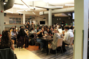 A crowd of students and community members sit around tables at the Observatory, an industrial-looking restaurant with support columns.