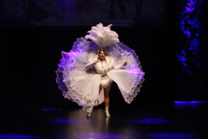 Kiki Coe is dressed in an intricate white feather and rhinestone garment as she stands in the middle of the stage.