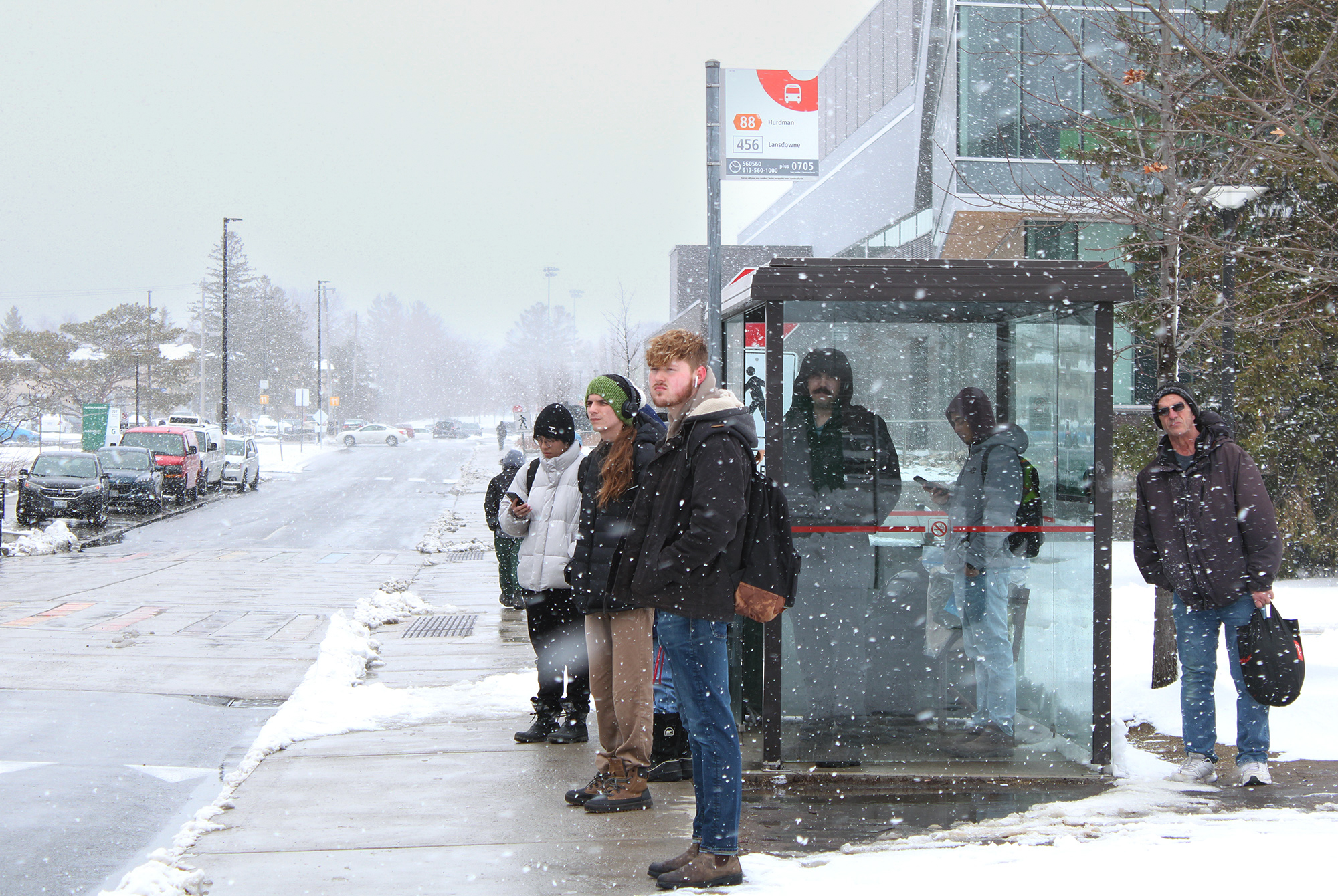 A group of students is waiting for the 88 bus under heavy snow in front of E-building at Algonquin College.