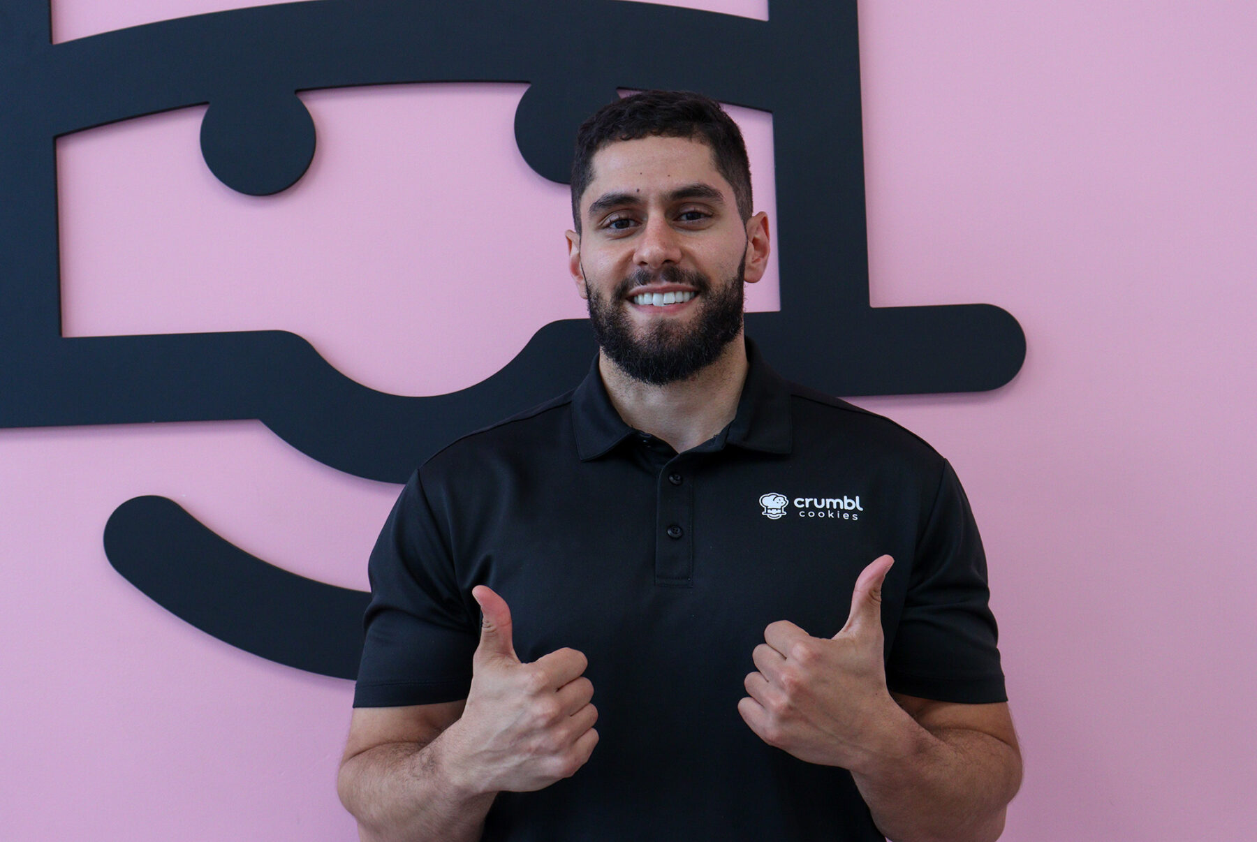 Owner Adam Al Hasan in front of the iconic pink wall and Crumbl logo.