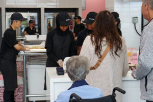 employees at Crumbl work serve customers with their orders. The open concept bakery is in full view with the ovens and lots of desserts being prepped.