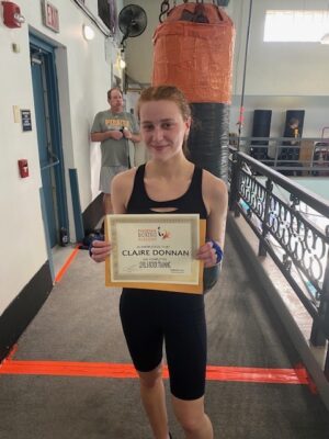 Claire Donnan, a white woman with red hair, stands in front of a punching bag in black biker shorts and a sports bra. Her hards are covered in blue hand wraps and she holds a certificate in her hands. She is visibly sweaty and red in the face.