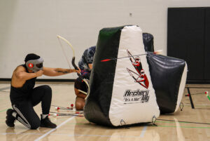 a player with a protective mask and a black athletic outfit is kneeling and drawing a bow, launching a foam-tipped arrow toward opponents.