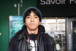 Gonzales is a tan man in a leather jacket, hat and scarf. He is looking at the camera in front of Savoir Fare