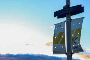 A signpost at the Fallowfield VIA Rail station featuring their branding. Below the sign is a banner displaying the slogan 'Love the Way.' The background reveals a blue sky with soft clouds illuminated by the warm glow of sunset.