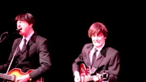 Close-up of two musicians on stage. The left-handed bassist and lead guitarist play side by side, focused on their instruments during a duet.
