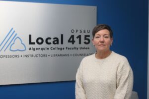 Tracy Henderson stands in front of the metal OPSEU 415 sign hanging in the union office in Algonquin Colleges A-building.